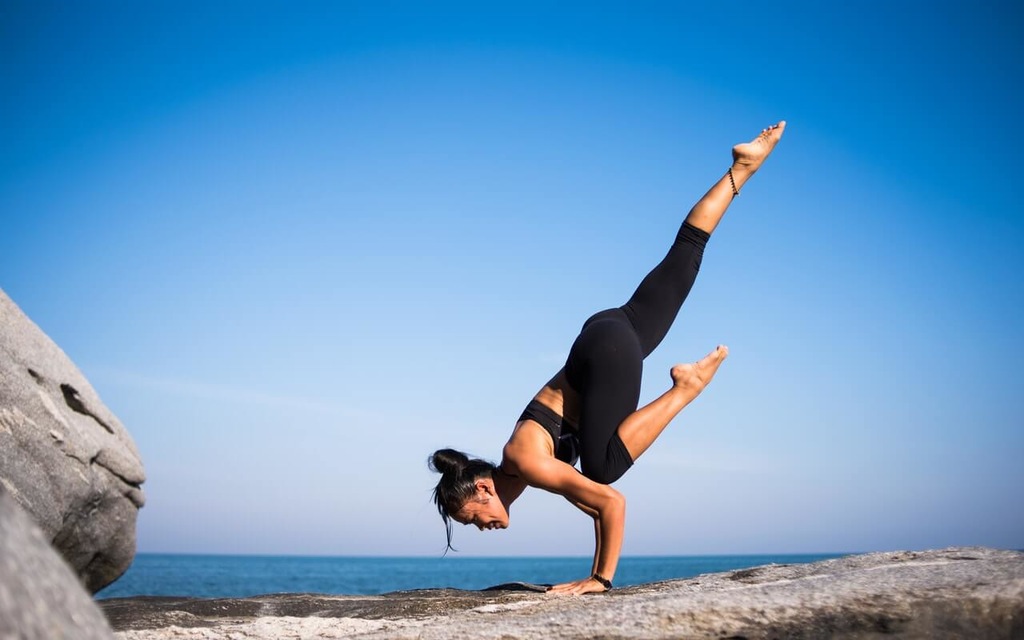 Mulher com roupas de ginástica fazendo posição de yoga numa pedra em frente ao mar